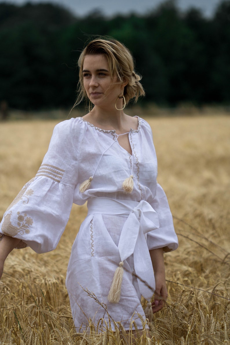 Puede incluir: Una mujer con un vestido blanco de lino con detalles bordados y borlas. El vestido tiene una cintura ce&ntilde;ida y una falda fluida. Est&aacute; de pie en un campo de hierba alta.