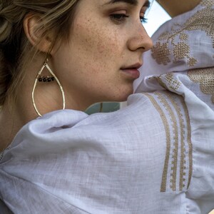 Puede incluir: Una mujer con pecas lleva una blusa de lino blanca con bordados dorados. Lleva un par de pendientes de gota dorados con cuentas marrones oscuras.