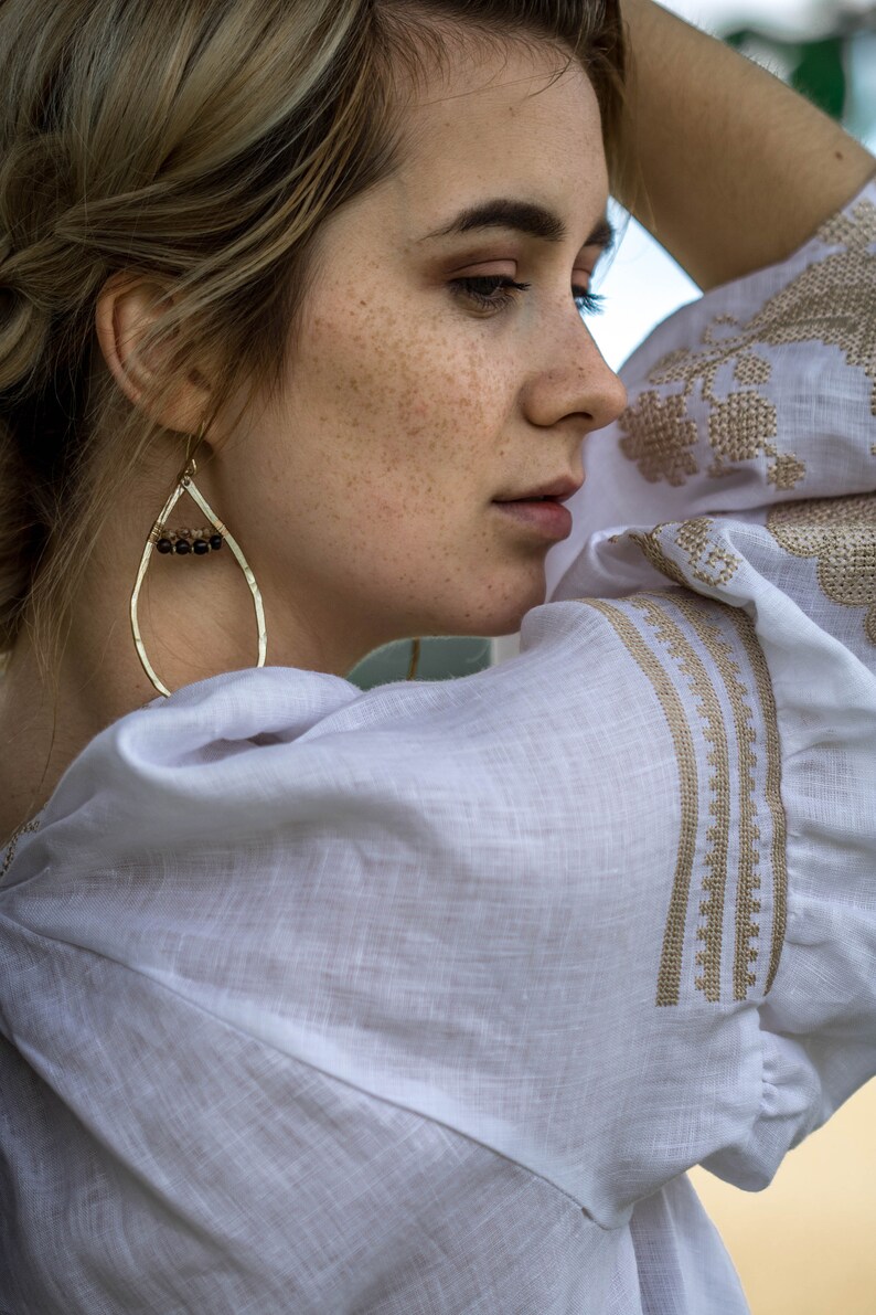 Puede incluir: Una mujer con pecas lleva una blusa de lino blanca con bordados dorados. Lleva un par de pendientes de gota dorados con cuentas marrones oscuras.