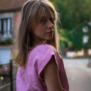 May include: A woman with long blonde hair wearing a pink linen top and a dark grey skirt. She is looking over her shoulder and smiling.