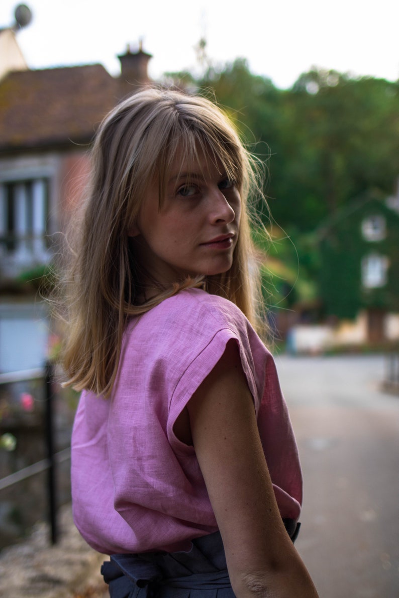 May include: A woman with long blonde hair wearing a pink linen top and a dark grey skirt. She is looking over her shoulder and smiling.