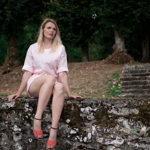 May include: A woman wearing a pink and white patterned short set sits on a stone wall. She is wearing orange sandals and looking to the right.