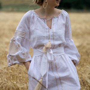 Puede incluir: Un vestido blanco de lino con detalles bordados y borlas. El vestido tiene una faja blanca atada a la cintura. La modelo est&aacute; de pie en un campo de trigo.