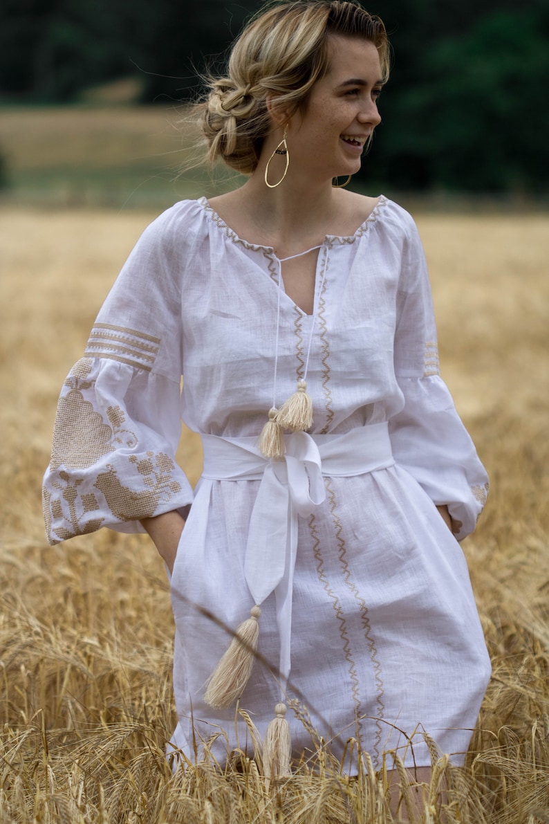 Puede incluir: Un vestido blanco de lino con detalles bordados y borlas. El vestido tiene una faja blanca atada a la cintura. La modelo est&aacute; de pie en un campo de trigo.