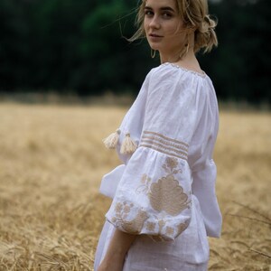 Puede incluir: Una mujer con un vestido de lino blanco bordado se encuentra en un campo de trigo alto y dorado. Est&aacute; mirando por encima del hombro con una sonrisa suave. El vestido tiene un corte holgado y fluido y presenta bordados delicados en color beige.