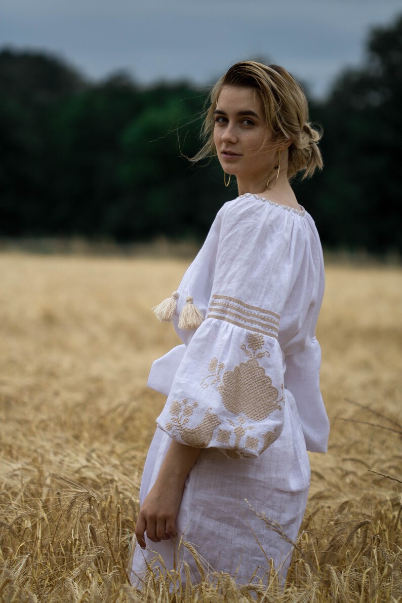 Puede incluir: Una mujer con un vestido de lino blanco bordado se encuentra en un campo de trigo alto y dorado. Est&aacute; mirando por encima del hombro con una sonrisa suave. El vestido tiene un corte holgado y fluido y presenta bordados delicados en color beige.