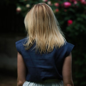 May include: A woman with long blonde hair is wearing a navy blue sleeveless top and a white skirt. She is standing in front of a blurred background of pink flowers.