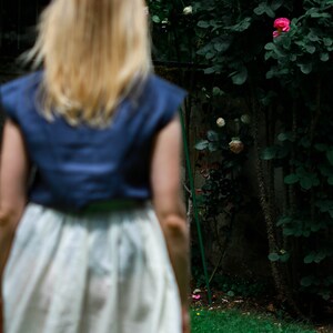 May include: A woman with long blonde hair wearing a blue sleeveless top and a white skirt stands in front of a lush green bush with pink roses.