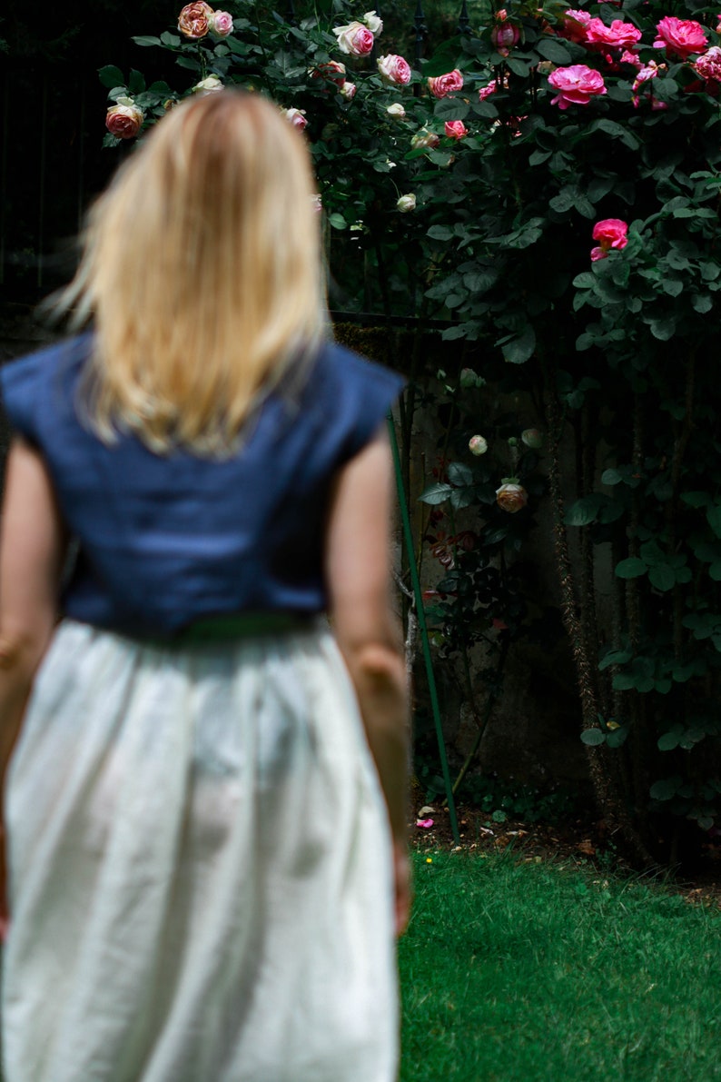 May include: A woman with long blonde hair wearing a blue sleeveless top and a white skirt stands in front of a lush green bush with pink roses.