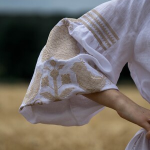 Puede incluir: Blusa blanca de lino con bordados de flores y p&aacute;jaros en hilo beige. La blusa tiene una manga abullonada y una cintura ajustada.