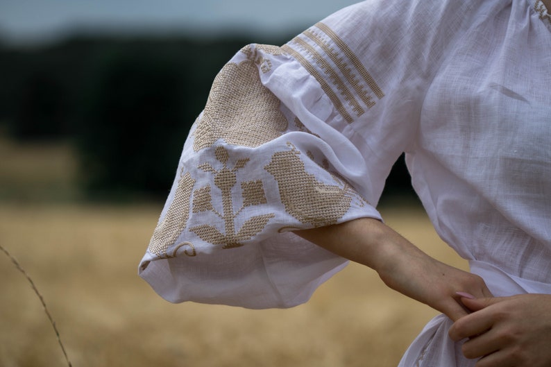 Puede incluir: Blusa blanca de lino con bordados de flores y p&aacute;jaros en hilo beige. La blusa tiene una manga abullonada y una cintura ajustada.
