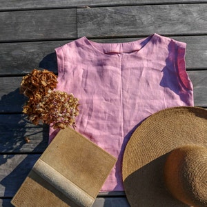 May include: A pink linen sleeveless top, a brown straw hat, a brown leather-bound book, and dried hydrangeas on a wooden surface.