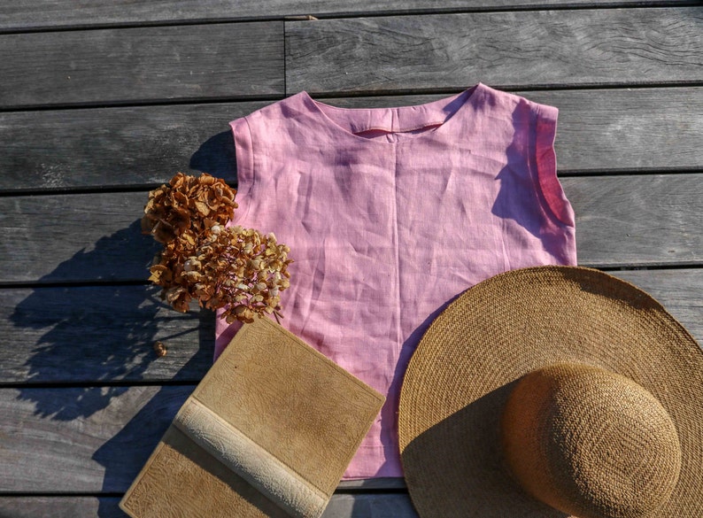 May include: A pink linen sleeveless top, a brown straw hat, a brown leather-bound book, and dried hydrangeas on a wooden surface.
