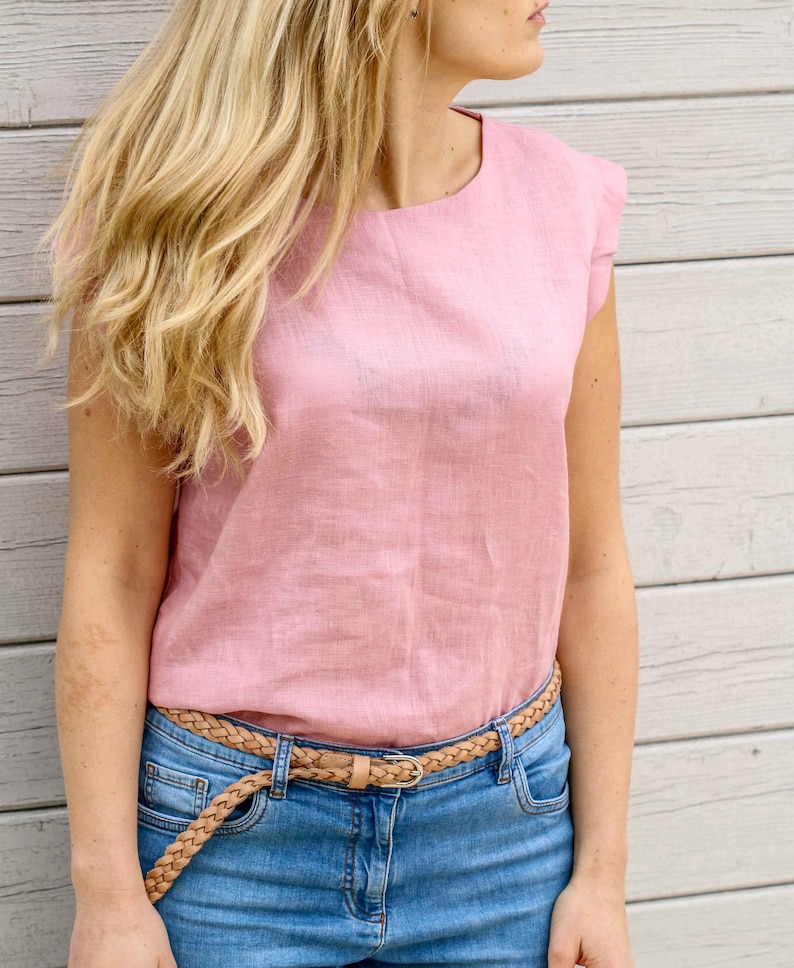 May include: A pink linen top with a round neckline and short sleeves. The top is worn with a brown braided belt and blue jeans.