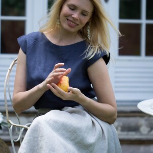 May include: A woman wearing a blue linen top and a light gray linen skirt sits on a white chair, holding a yellow pear in her hands.