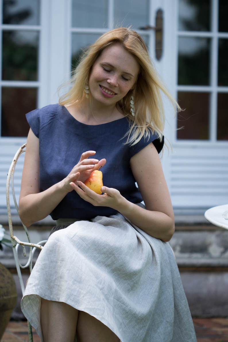 May include: A woman wearing a blue linen top and a light gray linen skirt sits on a white chair, holding a yellow pear in her hands.