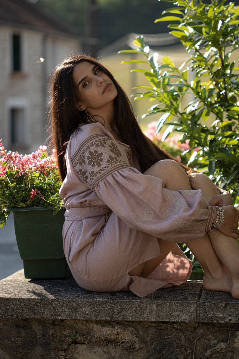 K&ouml;nnte beinhalten: Eine Frau in einem hellrosa Leinenkleid mit Stickereien sitzt auf einer Steinmauer. Sie hat langes dunkles Haar und tr&auml;gt einen goldenen Creolenohrring. Das Kleid ist locker geschnitten und perfekt f&uuml;r einen Sommertag.