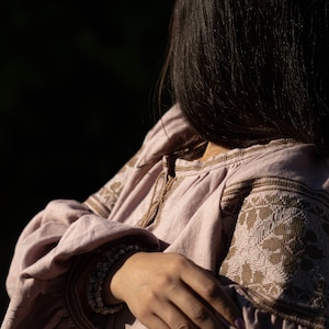 K&ouml;nnte beinhalten: Eine Frau tr&auml;gt eine hellrosa Leinenbluse mit braunen Stickereien. Die Bluse hat einen gerafften Ausschnitt und lange &Auml;rmel. Sie tr&auml;gt ein silbernes Armband am Handgelenk.