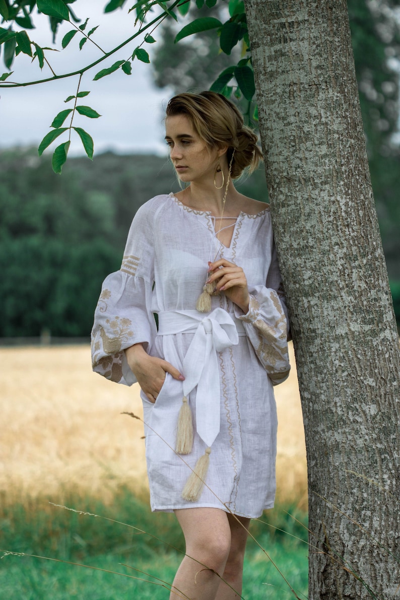 Puede incluir: Una mujer con un vestido blanco de lino con detalles bordados y un cintur&oacute;n blanco. El vestido tiene borlas en el cintur&oacute;n y las mangas. Est&aacute; de pie junto a un &aacute;rbol.