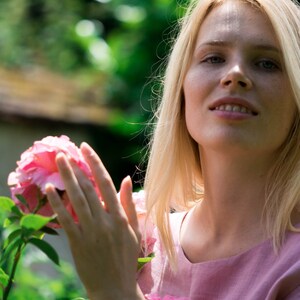 May include: A woman with blonde hair is standing in a garden, holding a pink rose in her hands. She is wearing a light pink dress.