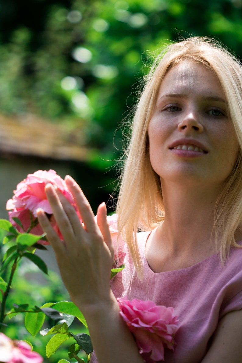 May include: A woman with blonde hair is standing in a garden, holding a pink rose in her hands. She is wearing a light pink dress.
