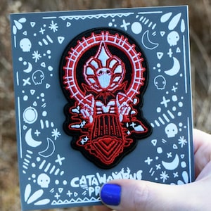 May include: A dark gray card with a decorative patch featuring a red and white design. The patch has a central figure with a halo-like ring. The card is decorated with white moons, stars, and skulls. The text "Catawampus Press" is visible.
