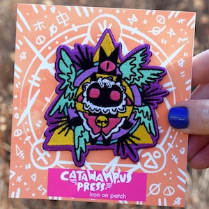 May include: A colorful iron-on patch featuring a cartoonish creature with a pink tongue, black eyes, and a yellow crown. The patch is on a peach-colored card with white symbols and the text "CATAWAMPUS PRESS iron on patch".