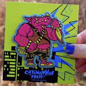 May include: A pink monster patch with a knife, on a neon green background with lightning bolts. The patch is an iron-on patch with the text "Catawampus Press". The patch is held by a hand with blue nail polish.