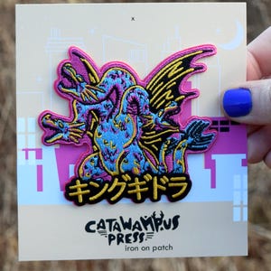 May include: An iron-on patch featuring a colorful, stylized illustration of a three-headed dragon with black wings and the Japanese text "キングギドラ". The patch is on a light pink and white card with the text "CATAWAMPUS PRESS iron on patch".