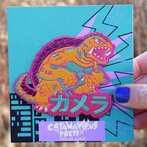 May include: An iron-on patch featuring a cartoon monster in shades of orange, yellow, and purple, with a turquoise background and Japanese text. The patch is rectangular with a black border and the text "CATAWAMPUS PRESS".