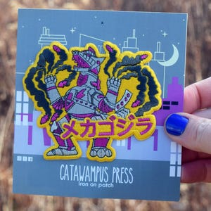 May include: Iron-on patch featuring a stylized monster design with the Japanese text "メゴジラ". The patch has a yellow border, a purple and white monster, and black smoke accents. The text "CATAWAMUS PRESS" is also visible.