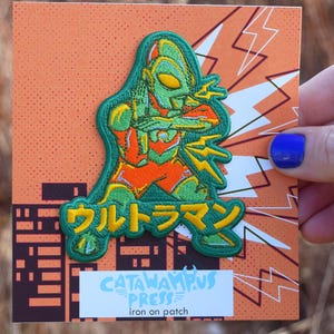 May include: An iron-on patch featuring a green and orange cartoon character with Japanese text. The patch is on a square orange background with a cityscape and lightning bolt design. The text "CATAMPUS PRESS" and "iron on patch" are also visible.