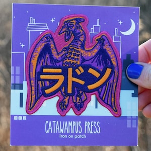 May include: A purple iron-on patch featuring a stylized illustration of a winged creature with Japanese text. The patch has orange and purple accents, with the text "CATAWAMUS PRESS" at the bottom.