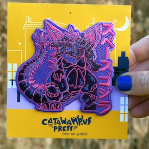 May include: A purple and black embroidered patch featuring a stylized monster design with Japanese text. The patch is on a yellow card with the text "Catawampus Press" and "iron on patch". The background is blurred.
