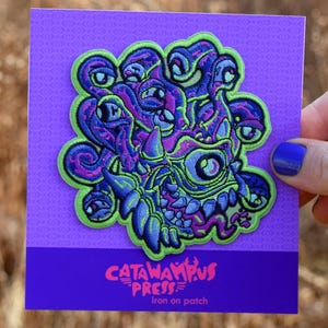 May include: A purple iron-on patch featuring a monster design with multiple eyes, teeth, and tentacles. The patch has green, blue, and purple accents. The text "CATAWAMPUS PRESS" is printed at the bottom of the purple card.