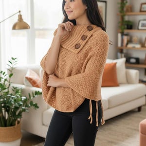 Peut inclure: Un poncho en crochet beige avec un col montant et trois gros boutons en bois. Le poncho a des manches évasées et des franges à l'ourlet. Le modèle porte un pantalon noir.