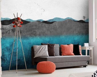 May include: A gray and blue abstract landscape wall mural with a gray couch and red ottoman in front of it. The couch has gray, black, and white patterned pillows and a red pillow.