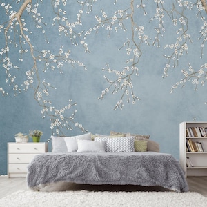May include: A bedroom scene featuring a light blue wall with a floral branch design. A bed with a gray fluffy comforter and pillows is in the foreground. A white dresser and bookshelf are also visible, creating a serene and inviting atmosphere.