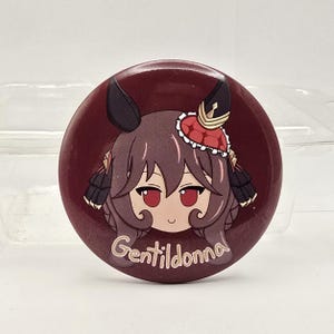 May include: A circular pinback button featuring a stylized anime-style character with brown hair, red eyes, and dark ears. The character wears a small red and white hat. The background is a deep maroon, and the text Gentildonna is printed in white.