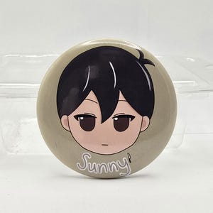 May include: A round button with a cartoon illustration of a character with black hair and brown eyes. The character's face is centered on a beige background. The word "Sunny!" is written in a cursive font at the bottom of the button.