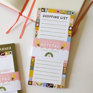 May include: A selection of stationery items, including a meal planner notepad, a shopping list notepad, and decorative tape rolls. The notepads feature floral and geometric designs with the text "MEAL PLANNER", "SHOPPING LIST", and "SPRING PATCHWORK NOTEPAD". An open notebook with a coral ribbon is also visible.