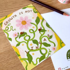 May include: A greeting card with the text "GROWTH IS NOT LINEAR" in a whimsical design. The card features a pink flower, green vines, and small snails. A blank white card and a colored pencil are in the background.