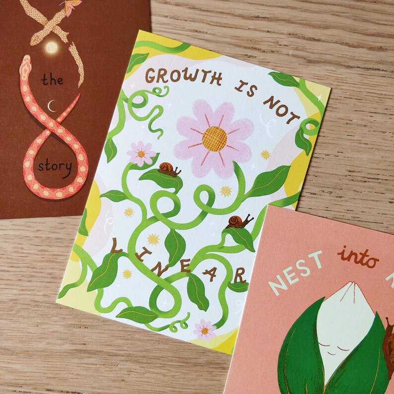 May include: Three art prints with illustrations and text. One print features a pink and orange snake in an infinity symbol shape with the words "the story". Another print has a pink flower, snails, and the words "Growth is not linear". The third print has the words "Nest into".