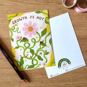 May include: A postcard with the text "GROWTH IS NOT LINEAR" in a whimsical design. The illustration features a pink flower, green vines, and small snails. A colored pencil and washi tape are also visible.