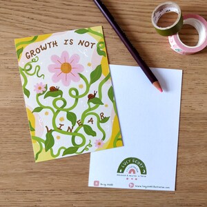 May include: A postcard with the text "GROWTH IS NOT LINEAR" and a floral design, alongside a blank card, a pencil, and decorative tape rolls. The postcard features a pink flower and green vines.