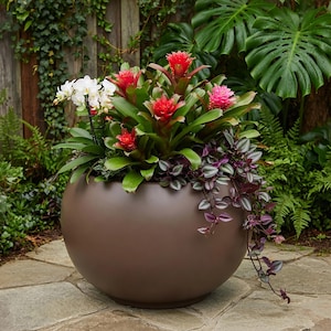 Op de afbeelding: Een grote, gladde, matbruine bolvormige plantenbak gevuld met diverse planten, waaronder witte orchideeën, verschillende rode en roze bromelia's met puntige groene bladeren, en een hangplant met kleine, paars en groen gevlekte bladeren.