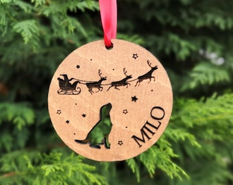 May include: A round wooden Christmas ornament with a red ribbon. The ornament features a laser-cut design of Santa's sleigh and reindeer, stars, and a dog silhouette. The name "MILO" is also laser-cut into the wood.