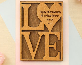 May include: A wooden card with the word "LOVE" in large letters. The letters are cut out and the background is a light brown wood. The card has a heart shape cut out in the center with the text "Happy 1st Anniversary All my love forever Mandy XXX" inside.
