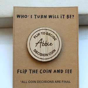 Puede incluir: Una moneda de madera con el texto "Flip to Decide Abbie Decision Coin" en una tarjeta marrón con el texto "Who's turn will it be? Flip the coin and see *All coin decisions are final"