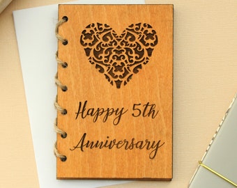 May include: A wooden card with a decorative heart design and the text "Happy 5th Anniversary". The card is tied with twine and has a white background.